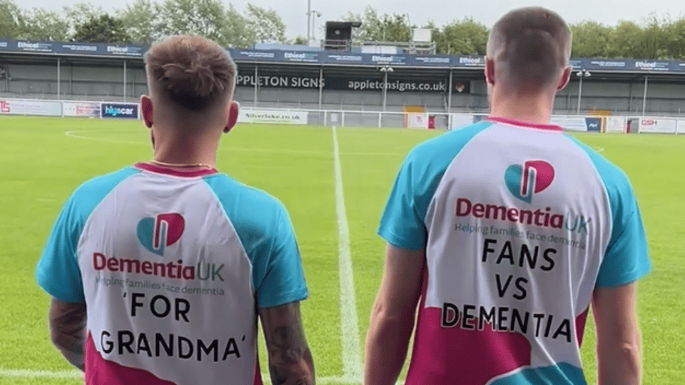 Two people stand on a football field wearing “Dementia UK” shirts; one reads “For Grandma,” the other reads “Fans vs Dementia.”.
