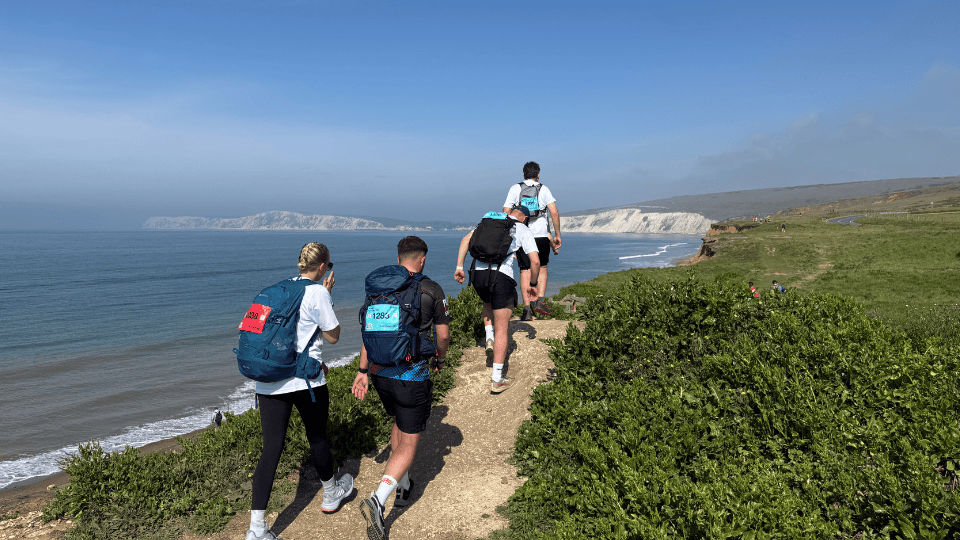 Four people wearing backpacks hike along a narrow coastal trail with cliffs and the sea visible in the background