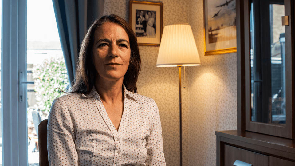 A woman with long brown hair and a patterned shirt sits indoors near a window, with framed photos, a lamp, and a cabinet in the background.