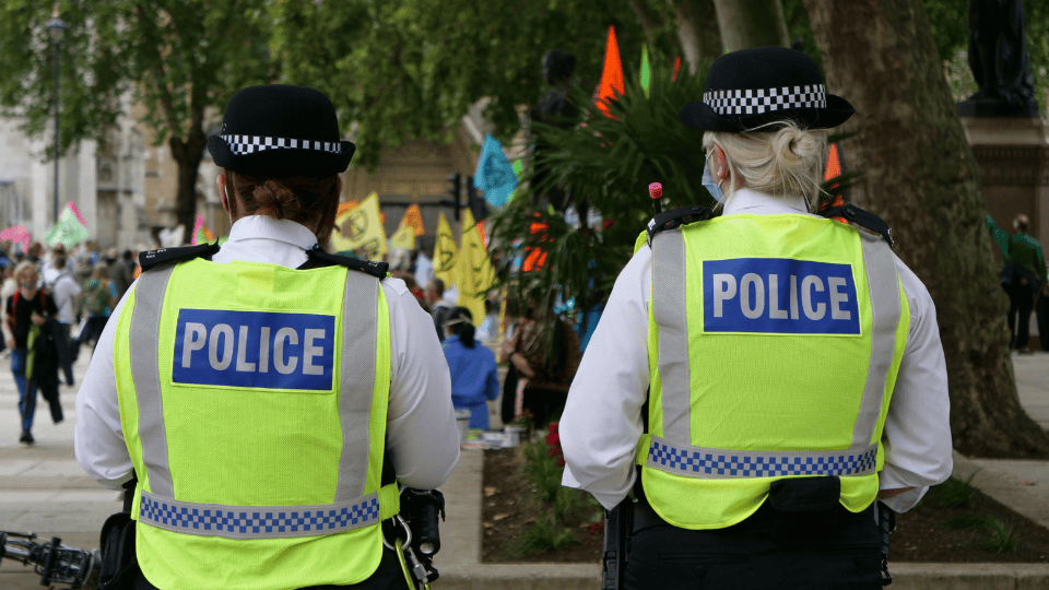 Two police officers wearing high-visibility vests with “POLICE” printed on the back stand in a park, facing a crowd.