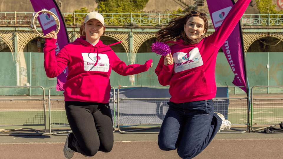 Two women in Dementia UK hoodies, one holding a tambourine, jump and smile outdoors in front of purple event banners and a fence.