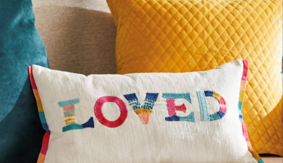 Dementia UK and Next collaboration cushion with the word Loved emboidered on it in rainbow colours.