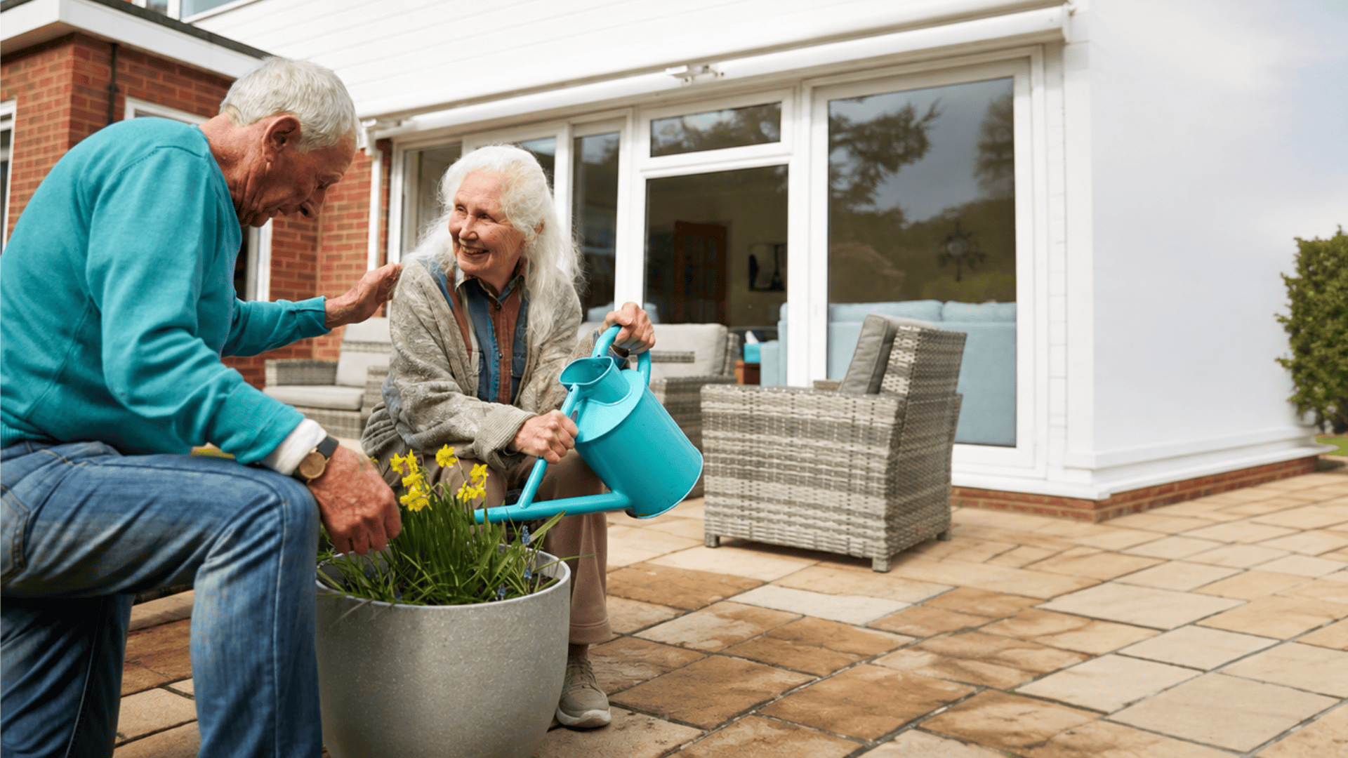 An elderly man and woman tend to potted flowers on a patio. The woman waters the plants with a blue watering can while the man smiles. Wicker chairs and a house are in the background.